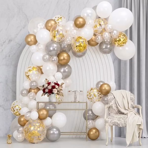 Arcada din baloane asamblata in culorile alb auriu chrome argintiu si cu confetti prezentata intr un decor luxos de eveniment cu fotoliu si flori