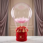 Aranjament elegant tip balon cu aer cald, format dintr-o cutie rosie cu trandafiri si un balon bobo transparent in interiorul caruia au fost introduse pene roz decorative.