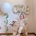 balon folie one argintiu 40x65 cm Decor festiv pentru prima aniversare a unui copil, evidentiind un balon din folie argintie in forma cuvantului cursiv ONE, fixat pe perete.