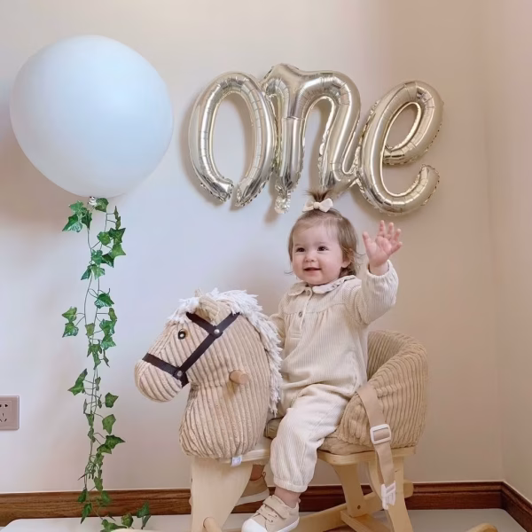 balon folie one argintiu 40x65 cm Decor festiv pentru prima aniversare a unui copil, evidentiind un balon din folie argintie in forma cuvantului cursiv ONE, fixat pe perete.