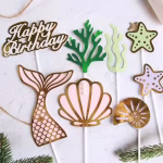 Set complet de 8 toppere pentru tort: coadă de sirenă, scoică cu perle, scoică spiralată, stele de mare, alge verzi și textul Happy Birthday auriu, montate pe bețe albe.