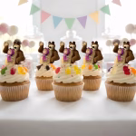 Set 10 Toppere Masha și Ursul pentru Tort și Brioșe – Decor Candy Bar Set 10 toppere decorative cu personajele Masha și Ursul înfipte în brioșe cu cremă albă pe o masă de candy bar aranjată festiv