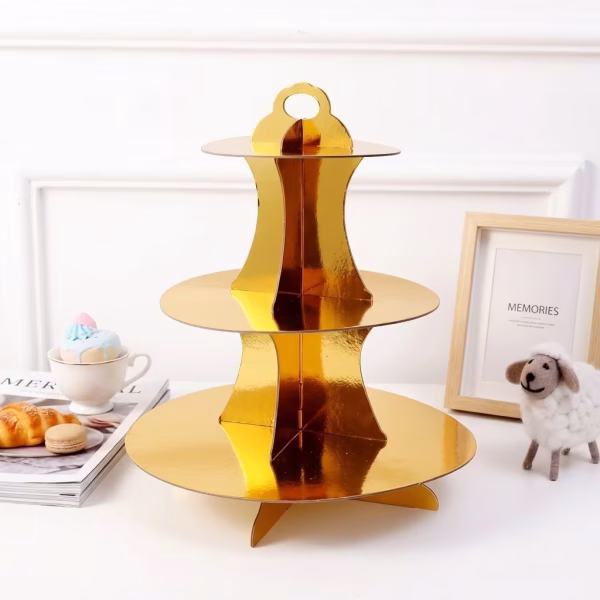 suport din carton pentru briose pe 3 niveluri auriu Suport din carton pentru briose pe 3 niveluri auriu lucios, asezat elegant pe o masa de Candy Bar alaturi de un desert si o rama foto.