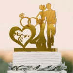 Topper de tort auriu din acril cu silueta mirilor inele si o inima cu textul Mr And Mrs asezat pe un tort alb cu frunze verzi