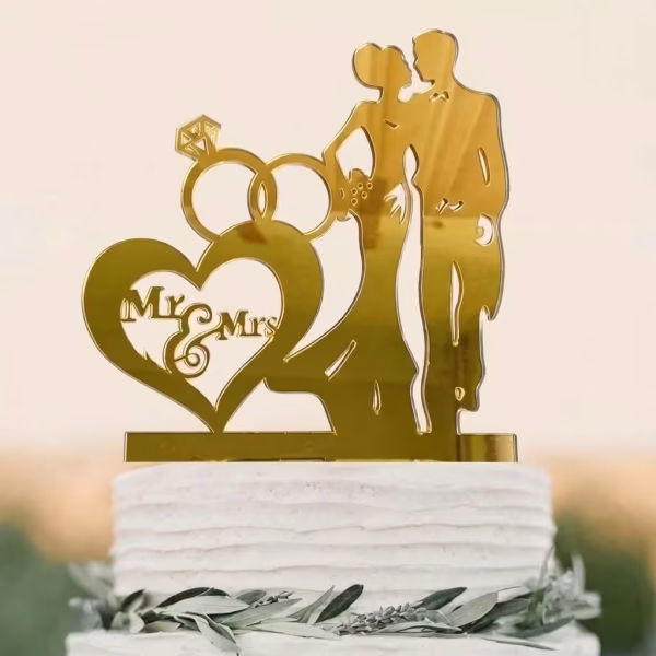 Topper de tort auriu din acril cu silueta mirilor inele si o inima cu textul Mr And Mrs asezat pe un tort alb cu frunze verzi