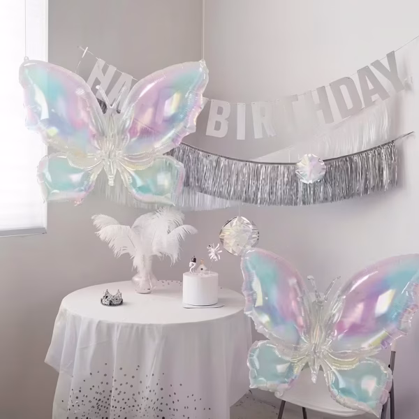 Decor de petrecere aniversara cu un balon folie fluture iridiscent gigant plutind deasupra unei mese cu tort alb accesorii argintii si fundal cu banner happy birthday