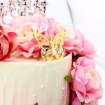 Topper de tort din plexiglass auriu in forma de fluture, cu o aripa decupata detaliat si o aripa solida gravata cu mesajul Happy Birthday, asezat printre trandafiri roz pe un tort