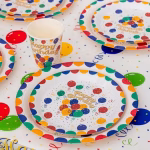 Decor de masa festiv pentru aniversare, cu farfurii din carton pentru tort (18 cm) asezate peste farfurii mari, insotite de pahare asortate cu model Happy Birthday.