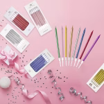 set 6 lumanari lungi pentru tort Set de 6 lumanari lungi si subtiri pentru tort, prezentate in mai multe cutii si culori lucioase pe un fundal roz festiv plin de confeti.