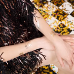 Femeie purtand o rochie festiva cu paiete negre, avand aplicate pe brate tatuaje temporare aurii si argintii cu pahare de sampanie si artificii.