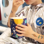 Bratul unui copil imbracat intr-un costum argintiu de astronaut, avand aplicate tatuaje temporare colorate cu un meteorit, un OZN si un extraterestru.