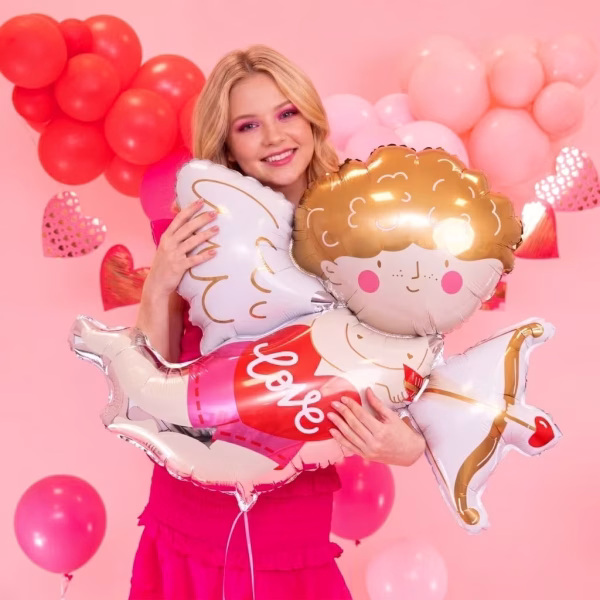 balon folie cupidon O tanara zambitoare tinand in brate un balon din folie gigant in forma de Cupidon cu arc si sageata, intr-un decor roz cu baloane inima.
