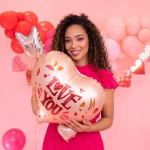 balon folie inima cu sageata O femeie zambitoare tinand in brate un balon din folie in forma de inima rose gold, strapuns de o sageata argintie, cu mesajul Love You.