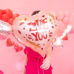 balon folie inima cu sageata O femeie zambitoare tinand in brate un balon din folie in forma de inima rose gold, strapuns de o sageata argintie, cu mesajul Love You.