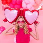 balon folie ochelari love O tanara zambitoare tinand in brate un balon din folie gigant in forma de ochelari cu inimi la o petrecere cu tematica roz.