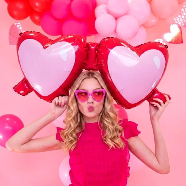balon folie ochelari love O tanara zambitoare tinand in brate un balon din folie gigant in forma de ochelari cu inimi la o petrecere cu tematica roz.
