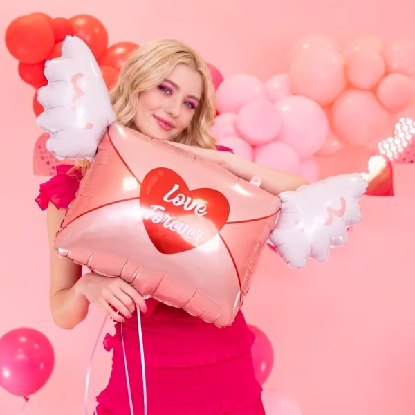 balon folie iubire pentru totdeauna O tanara blonda zambitoare tinand in brate un balon din folie in forma de plic roz cu aripi albe si o inima rosie cu textul Love Forever.