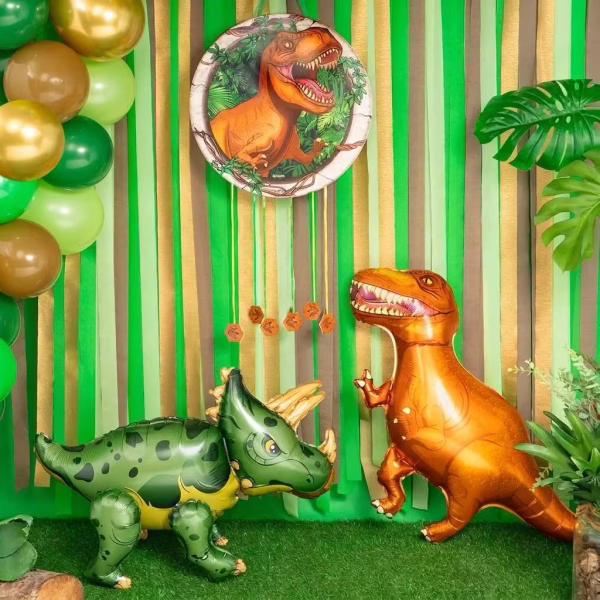 balon folie dinozaur airwalker Decor tematic de petrecere cu dinozauri, incluzand un balon folie triceratops verde si un t-rex portocaliu asezate pe iarba artificiala, inconjurate de un fundal de jungla.