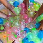 Mai multe maini de copii care iau baloane rotunde si colorate umplute cu apa dintr-un recipient transparent.