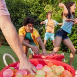 Adolescenti si tineri la o petrecere in curte, incarcandu-si mainile cu baloane colorate cu apa din doua galeti rosii mari.