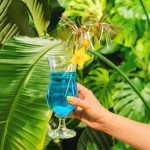 Pahar cu un cocktail albastru și o felie de fruct carambola, decorat elegant cu bețișoare tip artificii aurii și argintii.