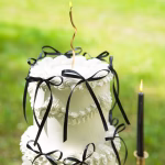 Set 10 Fundițe Negre Decor Tort Tort alb etajat, elegant, decorat festiv cu multiple fundițe negre din panglică subțire, așezat pe o masă rotundă acoperită cu o față de masă neagră.
