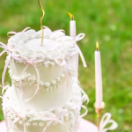 Set 10 Fundițe Roz Decor Tort Tort alb etajat, romantic, decorat festiv cu multiple fundițe roz pal din panglică subțire, așezat pe o masă rotundă într-un decor natural.