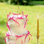 Set 10 Fundițe Magenta Decor Tort Tort alb etajat, elegant, decorat festiv cu multiple fundițe magenta din panglică subțire, așezat pe o masă rotundă acoperită cu o față de masă roz.