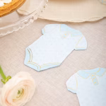 Servetele Forma Body Bebelus Bleu & Auriu Servetel decupat in forma de body de bebelus, asezat pe o fata de masa eleganta de in, langa o floare delicata si o farfurie pentru desert.
