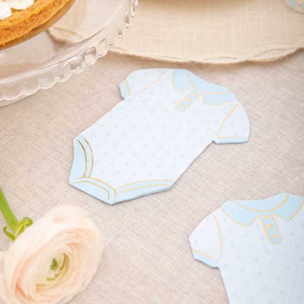 Servetele Forma Body Bebelus Bleu & Auriu Servetel decupat in forma de body de bebelus, asezat pe o fata de masa eleganta de in, langa o floare delicata si o farfurie pentru desert.