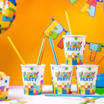Patru pahare din carton cu imprimeu Lego colorat si textul Block Party, asezate pe o masa festiva cu paie colorate.