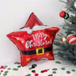 Balon Folie Craciun 45 cm Stea Mos Craciun Balon din folie rosie in forma de stea, decorat ca un costum de Mos Craciun cu mesajul Merry Christmas, asezat pe o masa cu confetti langa un brad impodobit.