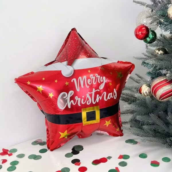 Balon Folie Craciun 45 cm Stea Mos Craciun Balon din folie rosie in forma de stea, decorat ca un costum de Mos Craciun cu mesajul Merry Christmas, asezat pe o masa cu confetti langa un brad impodobit.