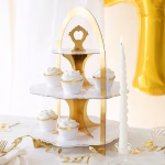 Suport etajat pentru prajituri alb cu auriu decorat elegant cu briose albe, asezat pe o masa festiva de petrecere alaturi de lumanari si ghirlande aurii.