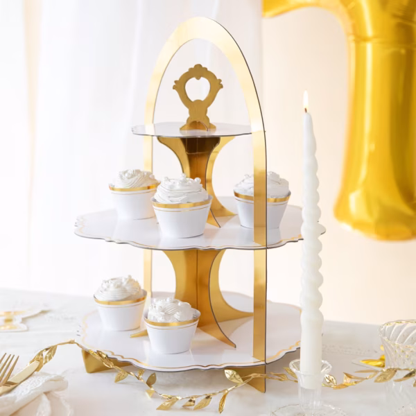 Suport etajat pentru prajituri alb cu auriu decorat elegant cu briose albe, asezat pe o masa festiva de petrecere alaturi de lumanari si ghirlande aurii.