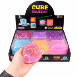 O mana sustinand un cub squishy transparent roz cu sclipici si paiete in interior, in fata unei cutii de prezentare cu 12 bucati asortate.