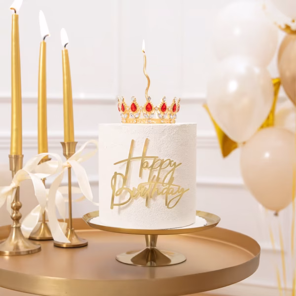 Topper Tort Coroniță Aurie cu Cristale Roșii 10cm Tort alb elegant așezat pe un suport auriu, decorat cu topperul coroniță cu cristale roșii, o lumânare spiralată și un mesaj frontal Happy Birthday auriu, înconjurat de baloane.