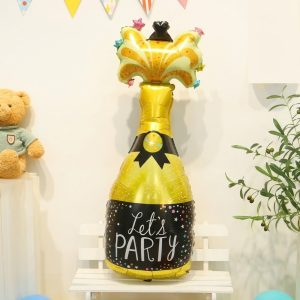 balon folie sticla de sampanie 88 cm Decor festiv cu un balon din folie gigant in forma de sticla de sampanie aurie desfacuta, asezat pe o bancuta alba.