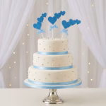 Set 3 Toppere Tort Inimi Bleu Tort alb etajat pe trei nivele, decorat cu panglici bleu la bază și perle argintii, având în vârf un set festiv de 3 toppere sub formă de cascadă de inimi bleu cu sclipici și fundițe.