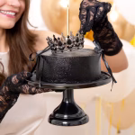 Topper Tort Coroniță Argintie cu Cristale Negre O persoană purtând mănuși negre din dantelă așază o coroniță metalică argintie cu cristale negre pe un tort elegant, complet negru, accesorizat cu panglici.