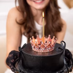 Topper Tort Coroniță Rose Gold cu Cristale O femeie zâmbind ține în mâini un tort negru elegant decorat cu o coroniță rose gold din metal și o lumânare lungă aprinsă.