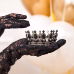 Coroniță argintie decorativă cu pietre negre sub formă de lacrimă și mici cristale rotunde, ținută cu delicatețe în palme acoperite de mănuși din dantelă neagră.