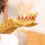 Coroniță aurie decorativă cu pietre roșii sub formă de lacrimă și cristale argintii, ținută în palme acoperite de mănuși elegante din satin auriu.