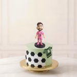 Figurină 3D topper tort cu Lionel Messi în echipamentul roz Inter Miami, așezată pe un tort cu decor de fotbal.