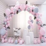 Kit Arcada Baloane Roz Si Gri Cu Suport Arcada din baloane roz pastel si gri decorata cu frunze artificiale asezata intr-un cadru festiv elegant cu scaun si suporturi albe.