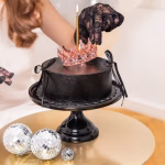 O mână purtând o mănușă din dantelă neagră ajustează o coroniță metalică rose gold cu cristale pe un tort negru mat, accesorizat cu panglici.