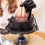O persoană cu mănuși din dantelă neagră aprinde o lumânare așezată în centrul unei coronițe rose gold aflate pe un tort negru.