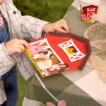 Mâinile unui copil ținând o farfurie din carton decupată în forma unui hambar roșu cu animale.