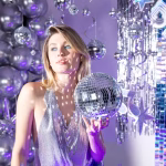 Femeie la o petrecere tematica tinand in mana un glob disco ale carui oglinzi reflecta raze luminoase pe fundal.