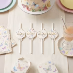 Masa pregatita pentru o petrecere de fetite cu tematica fluturi incluzand paie decorative pahare pastelate farfurii si baloane asortate